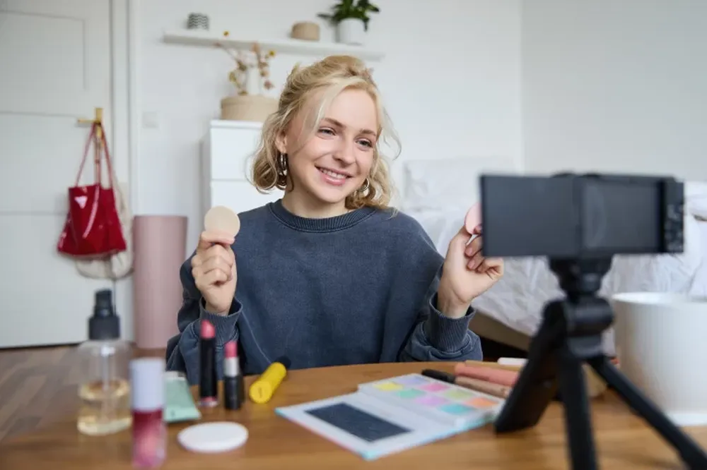 Portrait of young woman, beauty content creator, sitting in a room in front of digital camera, recording makeup tutorial vlog, showing cosmetic facial products.