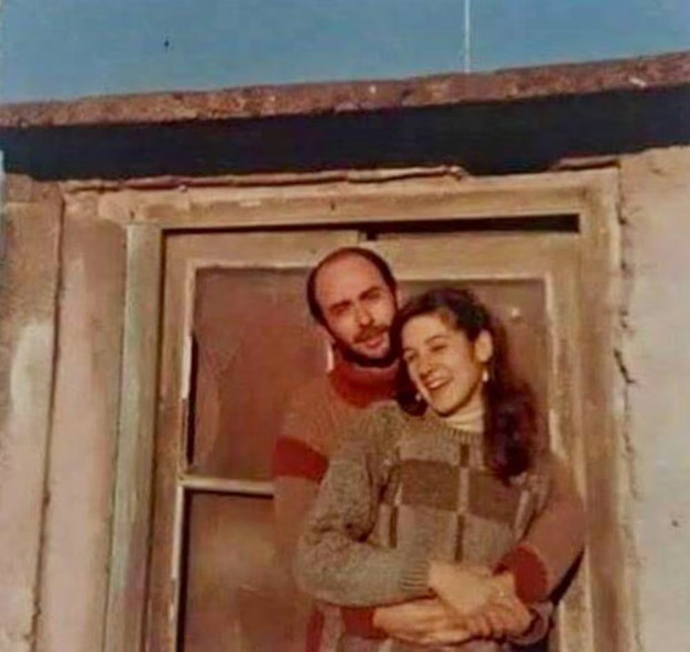 Indio Solari y Virginia sonríen en una tierna imagen de su juventud, abrazándose con cariño frente a una ventana.