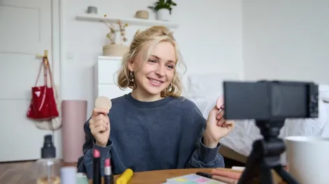 Portrait of young woman, beauty content creator, sitting in a room in front of digital camera, recording makeup tutorial vlog, showing cosmetic facial products.